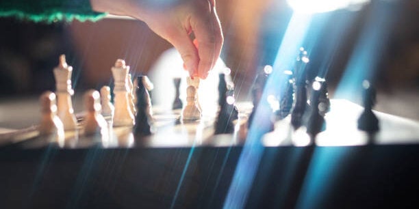 Close up of chess pieces on a chess board, sunlight streaming in a window behind with bright dramatic lens flare.  A childs hand picks up a piece to move it against their opponent.
